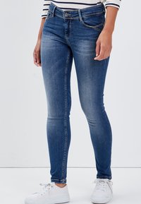 Dunkelblaue Skinny-Jeans aus Denim mit fünf Taschen, einem Knopf- und Reißverschluss sowie dezenten Used-Effekten an der Vorderseite. Weiße Sneaker runden den Look ab.