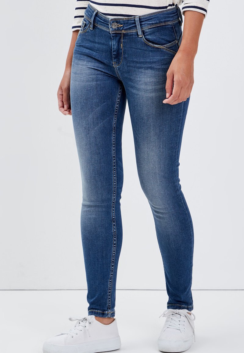 Dunkelblaue Skinny-Jeans aus Denim mit fünf Taschen, einem Knopf- und Reißverschluss sowie dezenten Used-Effekten an der Vorderseite. Weiße Sneaker runden den Look ab.