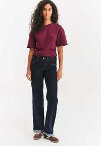 Žena s kudrnatými vlasy oblečená v vínovém tričku, tmavě modrých džínech s ohrnutými nohavicemi a hnědých teniskách, stojící na bílém pozadí.