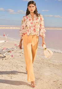 Blusa floreale in tonalità pastello con maniche a volant, abbinata a pantaloni a gamba larga color pesca e tacchi con cinturini, mentre tiene una borsa color crema.