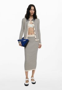 Set di cardigan e gonna a maglia rigata in crema con strisce blu. La modella porta una borsa geometrica blu e indossa scarpe bianche con dettagli neri.