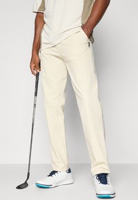 Beige golf pants featuring a relaxed fit, side pockets, and lightweight fabric paired with white and navy golf shoes. Golf club in hand.