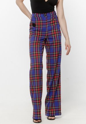 Kobieta ubrana w wysokie, niebiesko-czerwone spodnie w kratę o szerokich nogawkach oraz czarne sandały na obcasie z odkrytymi palcami, stojąca na tle jednolitego tła.