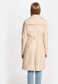 Beige trenchcoat met een getailleerd ontwerp, een ceintuur in de taille en lange mouwen. Heeft een achteropening en subtiele gouden hardwaredetails.