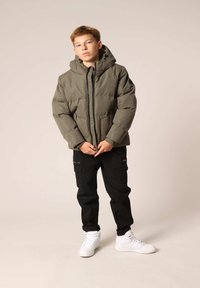 Veste olive verte rembourrée avec capuche, fermeture éclair et poches latérales, associée à un pantalon cargo noir et des baskets blanches. Texture lisse.