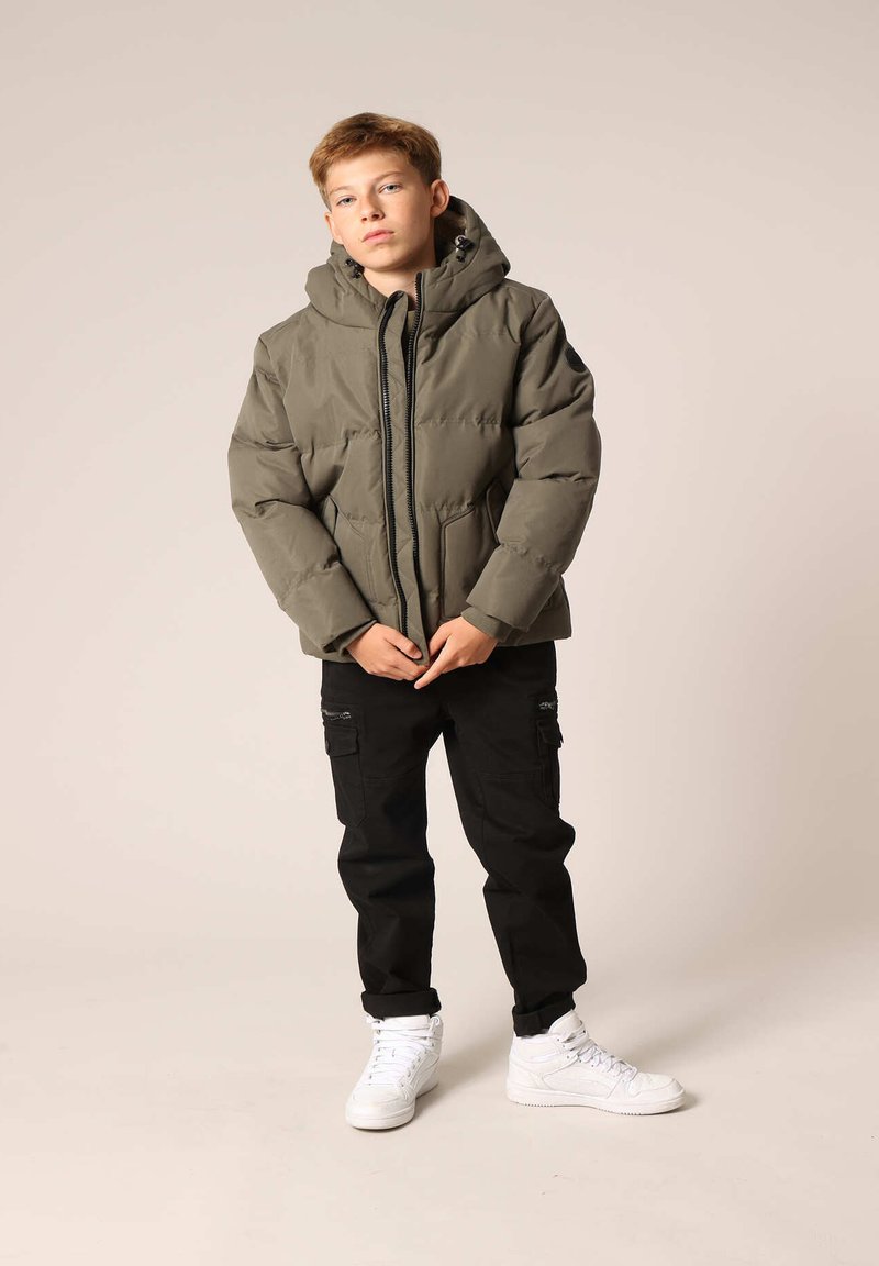 Veste olive verte rembourrée avec capuche, fermeture éclair et poches latérales, associée à un pantalon cargo noir et des baskets blanches. Texture lisse.