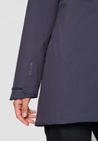 Marinblå vattentät jacka med en slät textur, justerbara manschetter och en diskret logotypdetalj på ärmen. Sidfickan är synlig.