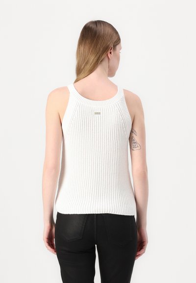 Débardeur sans manches blanc en maille côtelée avec un col rond et une texture légère, arborant une petite étiquette dans le haut du dos. Porté avec un pantalon noir.