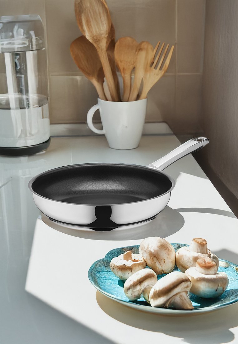 Stekpanna i rostfritt stål med svart non-stick insida, placerad bredvid en turkos tallrik med vita svampar på en vit bänkskiva.