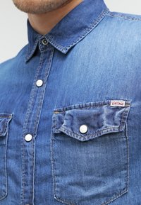 Koszula dżinsowa z klasycznym kołnierzykiem, srebrnymi zatrzaskami, dwiema kieszeniami na piersiach i vintage'ową metką. Materiał ma głęboki niebieski odcień.