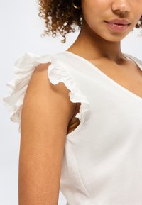 Γυναίκα που φορά ένα λευκό αμάνικο τοπ με βολάν στους ώμους και μικρό χρυσό κρίκο αυτιού, φωτογραφία κομμένη στο ύψος του στήθους και του βραχίονα.