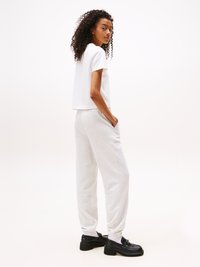 Jeune femme aux cheveux bouclés portant un t-shirt court blanc, un pantalon de survêtement gris clair, des chaussettes blanches et des mocassins noirs épais, se tenant de côté.