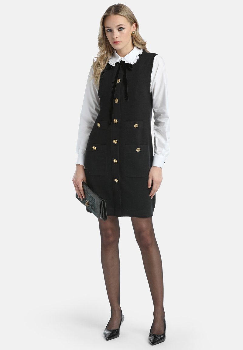 Donna che indossa un vestito senza maniche nero con bottoni dorati sopra una blusa bianca, tiene in mano una pochette nera e indossa tacchi neri e collant trasparenti.