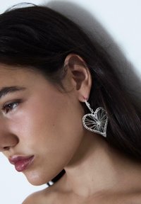 Primo piano di una donna con capelli scuri che indossa un grande orecchino argento a forma di cuore con dettagli intricati simili a una ragnatela.