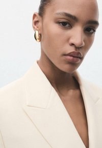 Oorbellen set in gouden metaal, met een ovale vorm. Het model draagt een lichtbeige blazer met een diepe V-halslijn. Glad textuur.