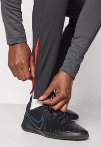Grå træningsbukser med en rød sideslids, parret med sorte sneakers med en grøn swoosh. Nærbillede af hænder, der justerer bukserne.