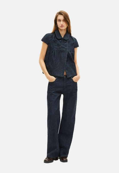 Completo in denim scuro composto da giacca corta con maniche a cappuccio e bottoni, abbinata a jeans a gamba larga con taglio dritto.
