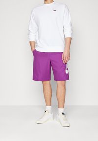 Fialové šortky s bílým logem Nike, spárované s bílou mikinou. Mikina má na hrudi malé červené logo. Na nohou má bílé tenisky.