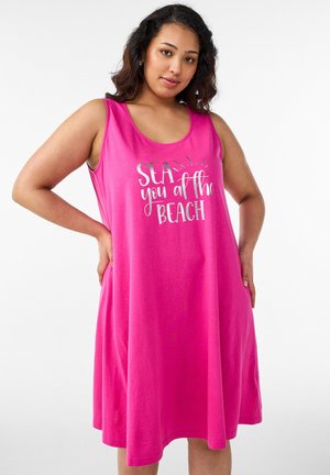Młoda kobieta o ciemnych włosach, ubrana w różową sukienkę bez rękawów z napisem „SEA you at the BEACH”, pozująca z rękami na biodrach.