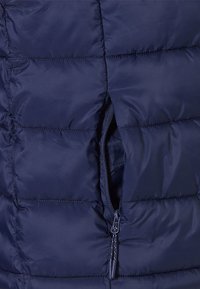 Marineblauwe pufferjack met een gewatteerde textuur en een ritszak aan de zijkant. Het materiaal lijkt lichtgewicht en waterafstotend.