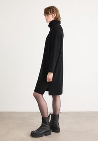 Zwarte coltrui jurk met lange mouwen en zijsplit, gecombineerd met chunky zwarte leren enkellaarsjes met een zijrits.