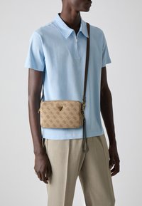 Smeđa crossbody torba s uzorkom, zlatnim logotipom, zatvaranjem na zip i odvojivim remenom. Nosi se preko svijetloplavog polo majice i bež hlača.