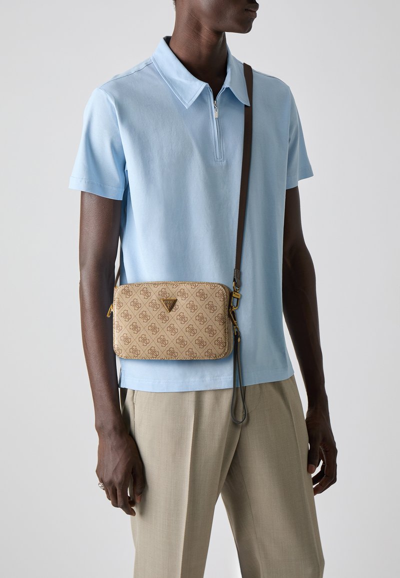 Smeđa crossbody torba s uzorkom, zlatnim logotipom, zatvaranjem na zip i odvojivim remenom. Nosi se preko svijetloplavog polo majice i bež hlača.