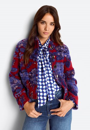 Woman with long brown hair wearing a red and purple textured jacket over a blue and white patterned tie-neck blouse and blue jeans, hands in pockets.
