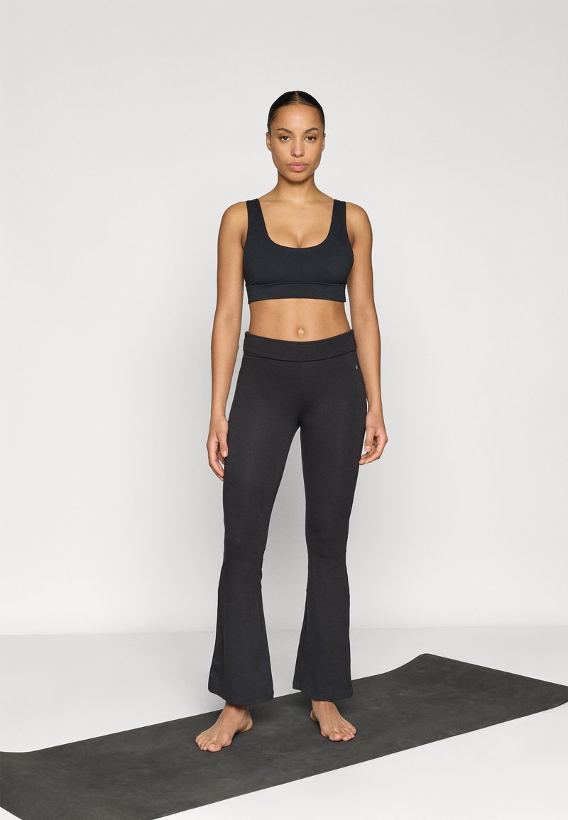 Soutien desportivo preto e leggings pretos evasê feitos de tecido elástico, usados por uma pessoa em pé sobre um tapete de yoga. Fundo neutro.