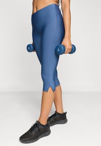 Μπλε αθλητικά κολάν με ψηλή μέση και σχιστό στρίφωμα, συνδυασμένα με μαύρα παπούτσια τένις από διχτυωτό ύφασμα. Το άτομο κρατά μπλε αλτήρες.