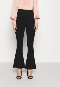 Zwarte flared broeken van rekbaar materiaal, met een hoge taille en een soepele, matte textuur. Gepaard met zwarte hoge hakken.