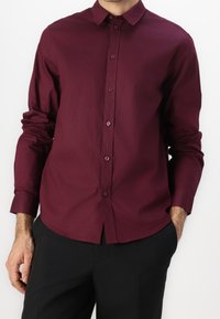 Bordowa koszula z długim rękawem, standardowym kołnierzykiem i zapięciem na guziki. Wykonana z gładkiego materiału, z pasującymi guzikami.