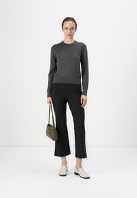 Grijze gebreide trui, zwarte slim-fit broek met een subtiele flare, en lichtgrijze instap schoenen. Kleine olijfgroene tas met een gestructureerde afwerking.