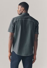 Camicia a maniche corte grigio scuro con impunture a contrasto, colletto alla coreana con bottoni e orlo leggermente arrotondato. Tessuto con trama visibile.