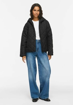 Donna con giacca nera trapuntata con cappuccio, camicia bianca, jeans larghi blu e scarpe nere, in piedi davanti a uno sfondo chiaro e semplice.
