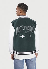 Chaqueta bomber verde con mangas blancas, que presenta un gran gráfico en la parte posterior con texto y diseños celestiales. Puños y bajo acanalados.