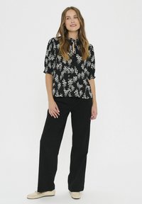 Blusa nera con motivo floreale bianco, scollatura rotonda con apertura a chiave, e maniche corte a sbuffo. Abbinata a pantaloni neri a vita alta e scarpe color sabbia.