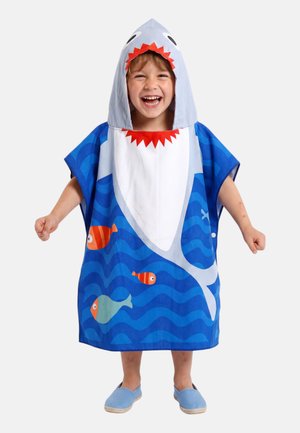 Enfant souriant portant une serviette à capuche bleue sur le thème du requin avec des motifs de poissons, debout avec les poings légèrement serrés et les pieds dans des chaussures bleues sans lacets.
