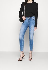 Frau trägt hellblaue Skinny Jeans, ein schwarzes Langarmoberteil, schwarze High-Heels und hält eine schwarze Umhängetasche mit Kette.