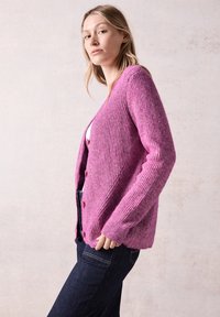 Ribbad rosa kardigan med långa ärmar och v-ringning, med knäppning och avslappnad passform. Bärs över en vit topp och mörka jeans.