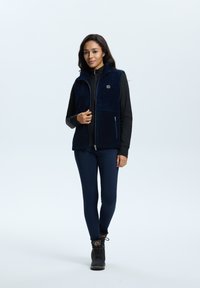 Marineblaue Fleecepulloverweste mit Reißverschluss, hohem Kragen und Logodetail. Kombiniert mit einem schwarzen Rollkragenpullover, marineblauen Leggings und schwarzen Stiefeln.