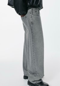 Weit geschnittene graue Jeans aus Denim mit strukturierter Oberfläche, ausgestattet mit Vordertaschen und einem Markenpatch auf der Gesäßtasche. Kombiniert mit schwarzen Stiefeln.