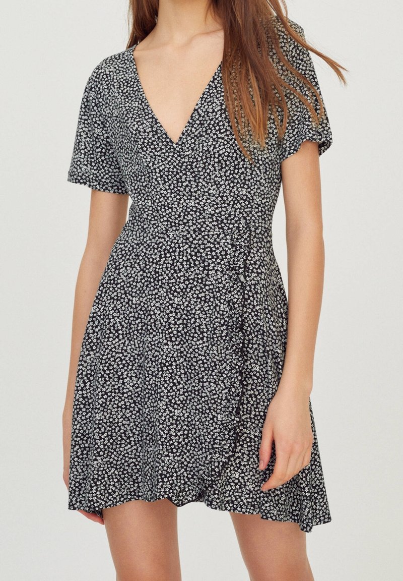 Femme portant une robe portefeuille noire à motifs floraux, manches courtes et décolleté en V, debout devant un fond clair uni.