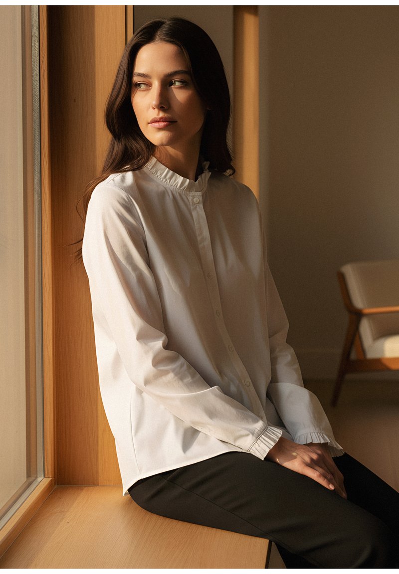 Vit skjorta med knappar och rynkad krage, kombinerad med svarta byxor. Mjuk tyg och avslappnad passform, sittande på en trälist vid ett fönster.