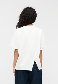 Λευκό βαμβακερό t-shirt με χαλαρή εφαρμογή, κοντά μανίκια και χαρακτηριστικό σχέδιο με σχίσιμο στην πλάτη. Η υφή φαίνεται λεία και καθημερινή.