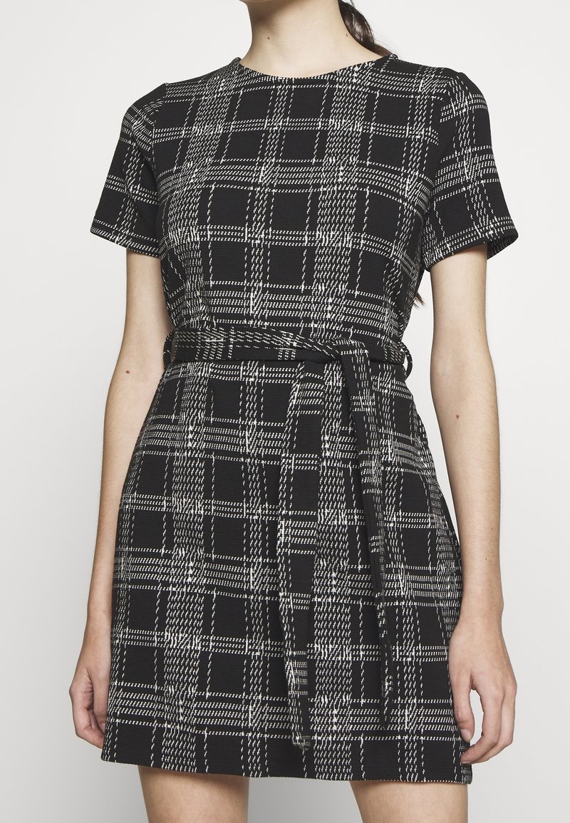 Rutig klänning i svart och vitt med korta ärmar, åtsittande midja och bältesdetalj. Tillverkad av textil med struktur, med rund halsringning.