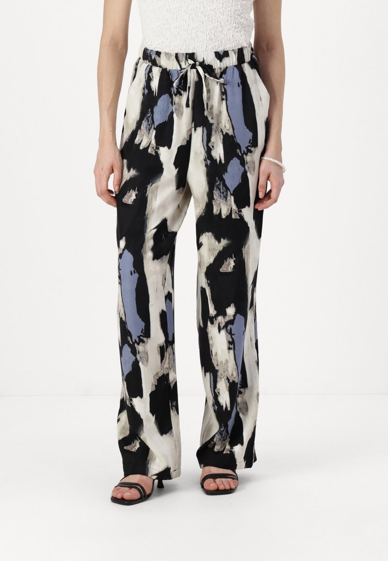 Femme portant un pantalon ample taille haute avec un imprimé abstrait noir, blanc et bleu, associé à des sandales à talons noires et un haut blanc.