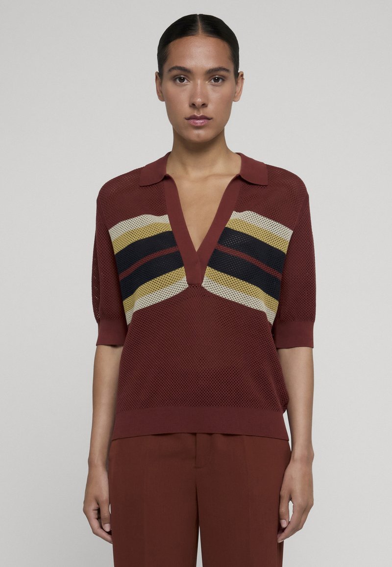Femme portant un top en maille résille couleur rouille avec un décolleté en V profond et des rayures chevrons multicolores, assorti à un pantalon, debout devant un fond uni.