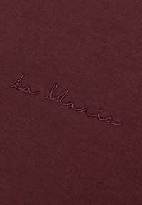 Bordowa tkanina o gładkiej fakturze z haftowanym napisem "La Mania" w kontrastującym kolorze, umieszczonym po przekątnej.