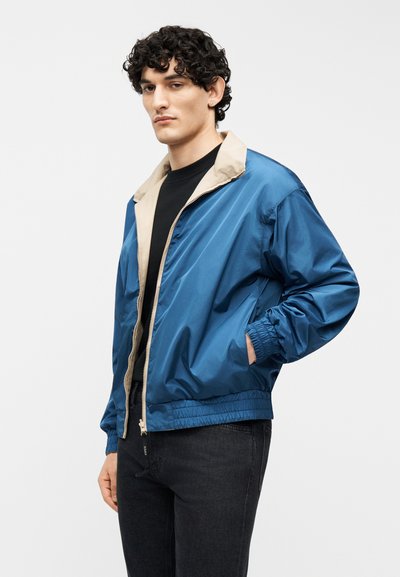 Νεαρός με σγουρά μαλλιά φορώντας μπλε μπουφάν με φερμουάρ και μπεζ γιακά πάνω από μαύρο πουκάμισο και μαύρο τζιν, με το χέρι στην τσέπη, ουδέτερο φόντο.